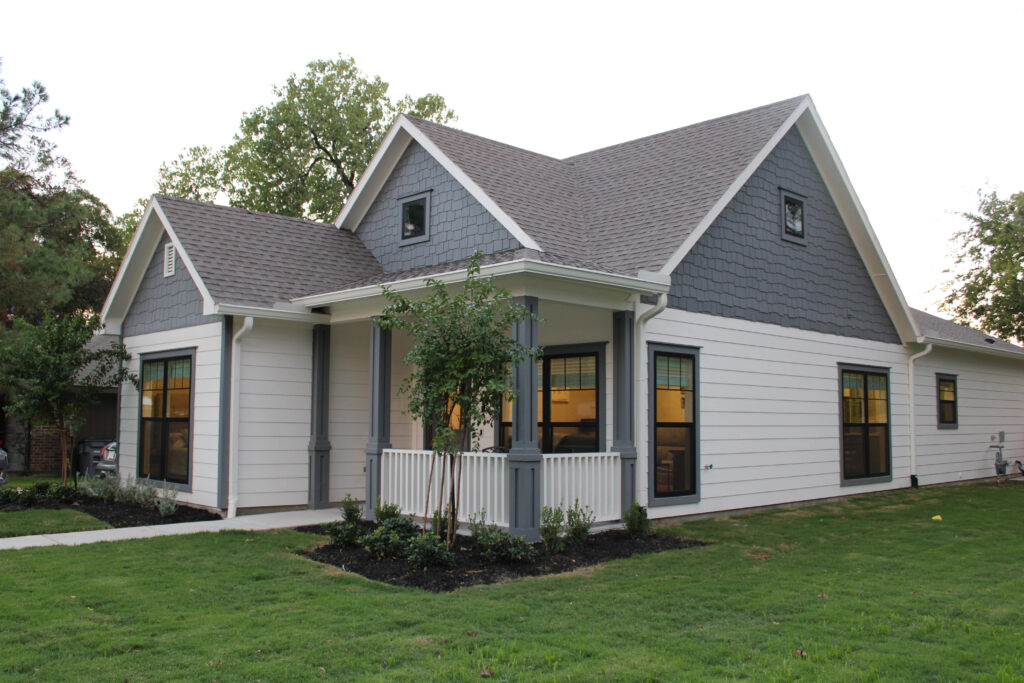 Angled view of a craftsman-style house with gray shingle accents, white siding, dark trim, and a covered front porch. The yard is freshly landscaped with green grass and young shrubs.