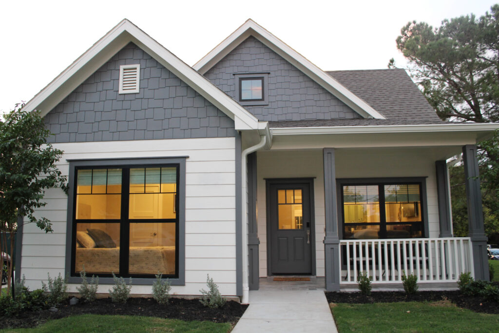 Front view of a modern craftsman-style home with gray and white siding, dark trim, and a small covered porch. Large front windows and neatly landscaped flower beds.